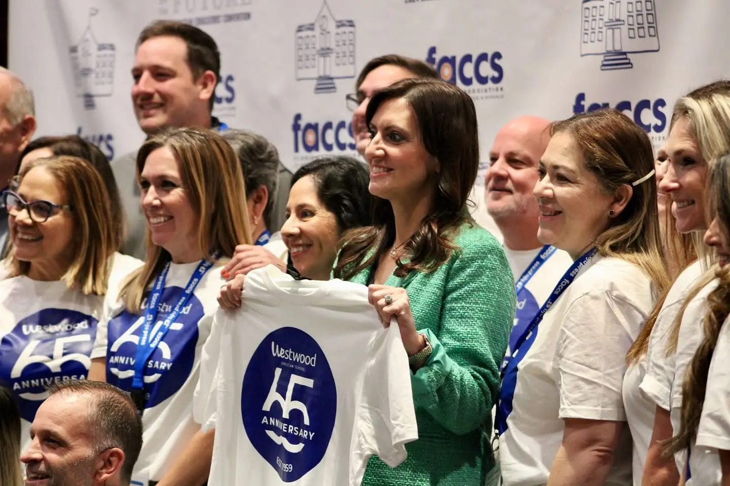 A group of educators holding up a school anniversary shirt at the FACCS backdrop.
