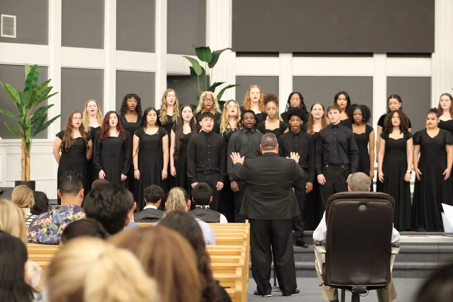 A large high-school choir in black performance dress singing on stage as their conductor directs.