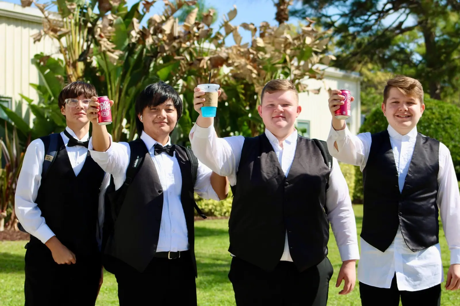 Four high-school students in performance vests and bow ties smiling and raising their drinks together outside the venue.