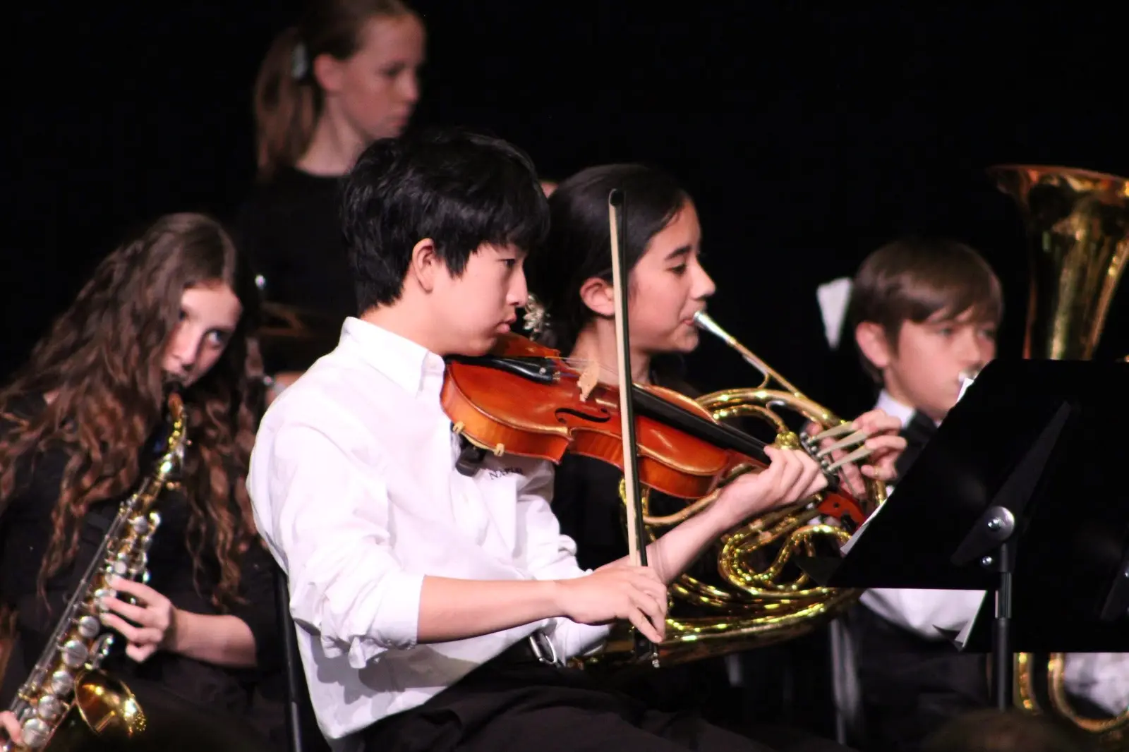 Student cellists and bassists mid-performance on stage, bows raised.