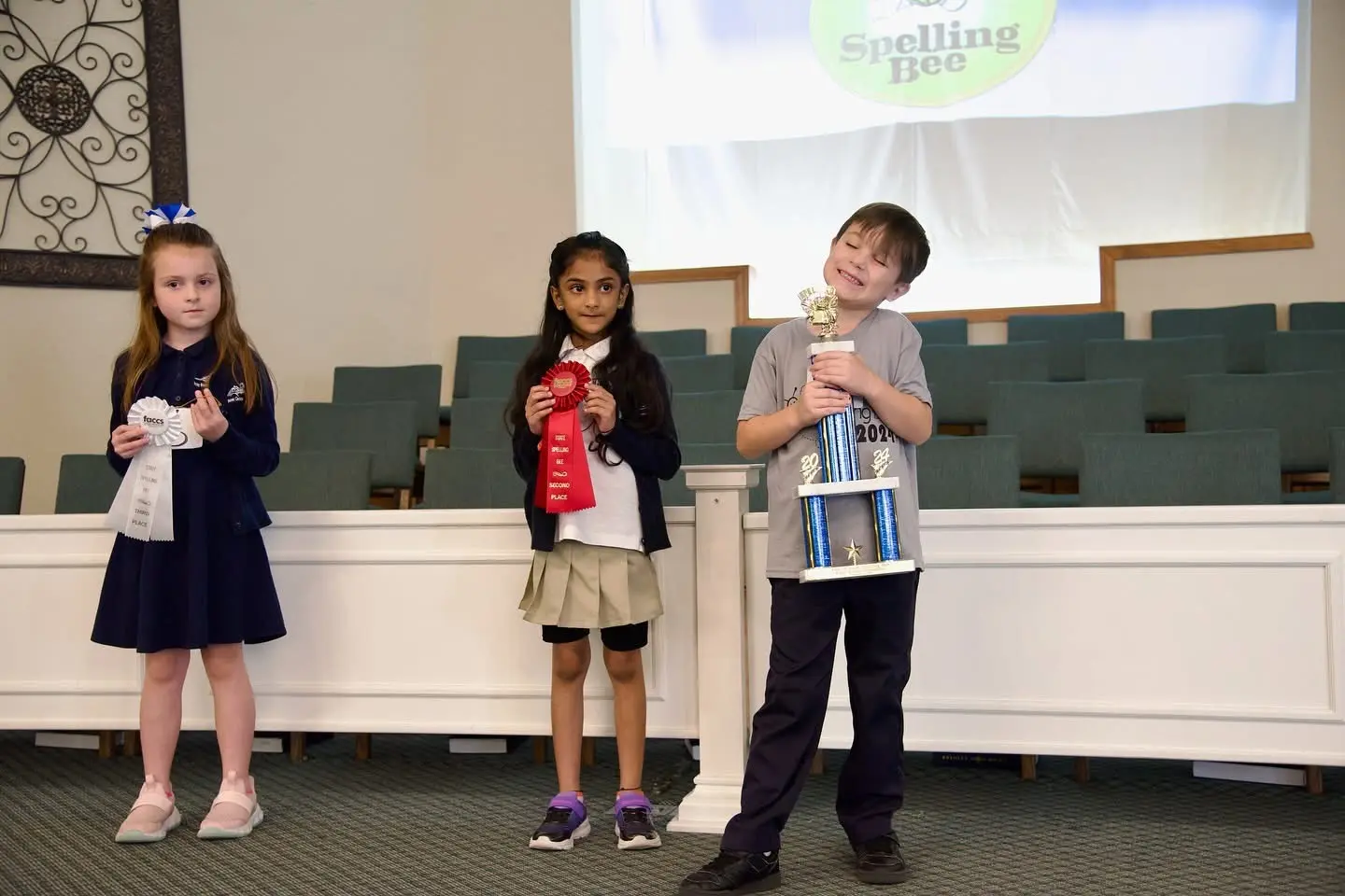 Three young spellers holding trophies and ribbons in front of the FACCS Spelling Bee banner.